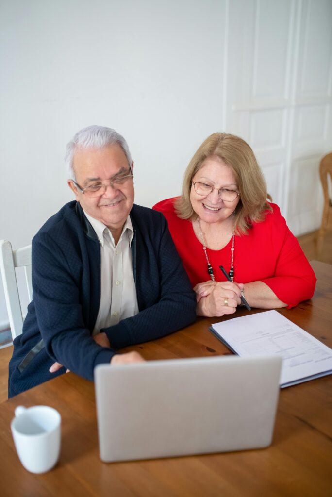 pexels-photo-8439751-8439751 Senior couple sitting at a table, smiling while using a laptop indoors. Perfect for tech and lifestyle themes.